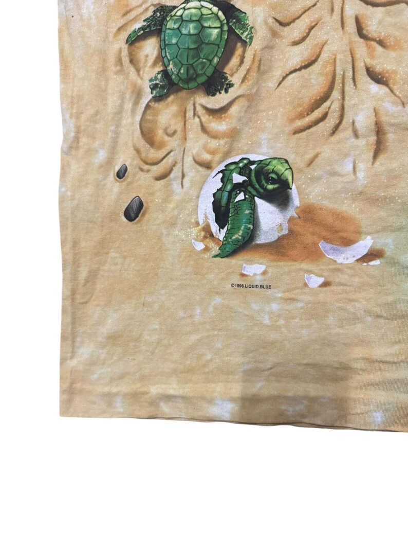 May include: A tan t-shirt featuring a detailed graphic of sea turtles. One turtle is emerging from an egg, while another is depicted above. The design includes sand-like textures and the text "&copy;1996 LIQUID BLUE".