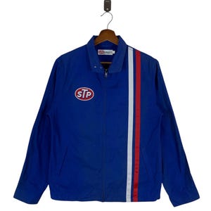 Op de afbeelding: Blauw jack met ritssluiting met het STP-logo op de borst en een verticale rood-wit-rode streep aan de rechterkant. De jas hangt aan een houten hanger. Klassieke racestijl.