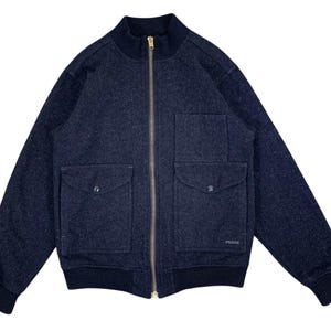 Puede incluir: Chaqueta bomber de lana azul marino con cremallera dorada y dos bolsillos delanteros. La chaqueta tiene cuello, pu&ntilde;os y dobladillo de canal&eacute;. El logotipo de Filson es visible en la parte inferior derecha.