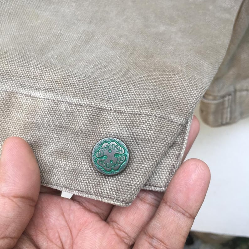 K&ouml;nnte beinhalten: Nahaufnahme einer braunen Canvasjacke mit einem gr&uuml;nen und silbernen Knopf, der ein vierbl&auml;ttriges Kleeblatt und den Text "C.K.O.S.H. F.C.O.S.H." zeigt.