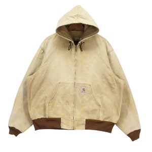 May include: A vintage Carhartt work jacket in a light beige color with a brown hood and brown ribbed cuffs. The jacket has a zipper closure and a patch pocket on the chest with the Carhartt logo.