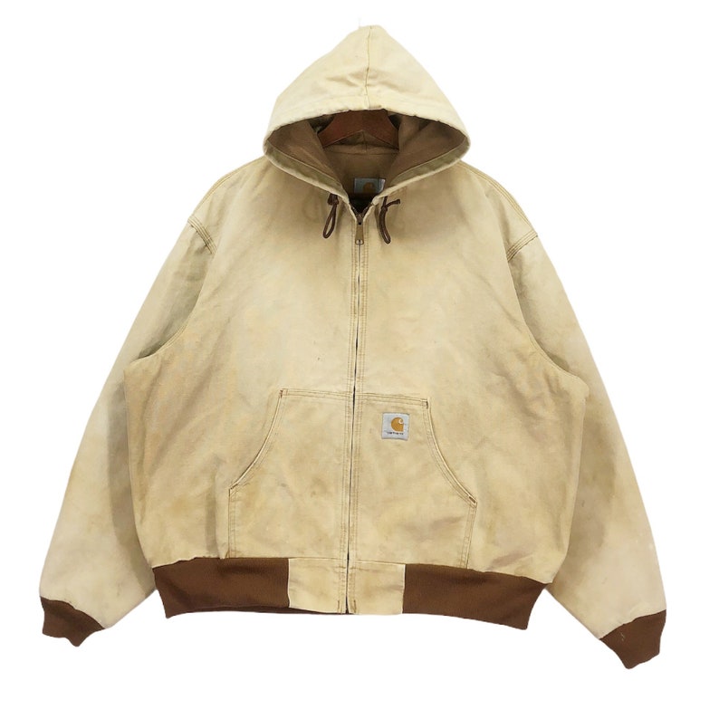 May include: A vintage Carhartt work jacket in a light beige color with a brown hood and brown ribbed cuffs. The jacket has a zipper closure and a patch pocket on the chest with the Carhartt logo.