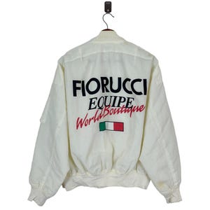 May include: White bomber jacket with a black and red "FIORUCCI EQUIPE World Boutique" logo and an Italian flag on the back.