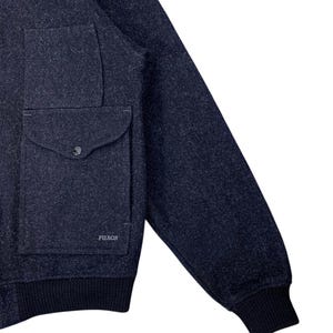 Puede incluir: Chaqueta de lana azul marino con un bolsillo grande y cierre de bot&oacute;n. La chaqueta tiene pu&ntilde;os y dobladillo de punto acanalado. El nombre de la marca "FILSON" es visible en el bolsillo. Una prenda de abrigo cl&aacute;sica.