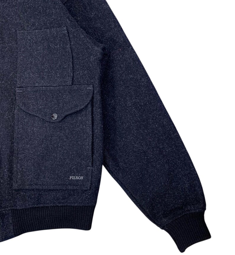 Puede incluir: Chaqueta de lana azul marino con un bolsillo grande y cierre de bot&oacute;n. La chaqueta tiene pu&ntilde;os y dobladillo de punto acanalado. El nombre de la marca "FILSON" es visible en el bolsillo. Una prenda de abrigo cl&aacute;sica.