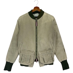 Puede incluir: Chaqueta bomber de color beige con cuello y puños verdes. La chaqueta tiene dos bolsillos delanteros grandes y una cremallera completa. El tirador de la cremallera tiene texto rojo y blanco. La chaqueta está colgada de una percha de madera.