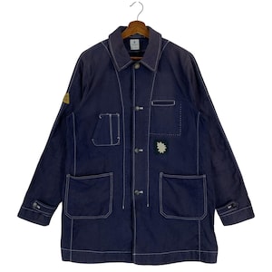 May include: A dark navy blue chore coat featuring contrasting white stitching, a pointed collar, and a button-front closure. The jacket includes two large lower patch pockets, a chest pocket, a small sleeve patch, and adjustable buttoned cuffs.