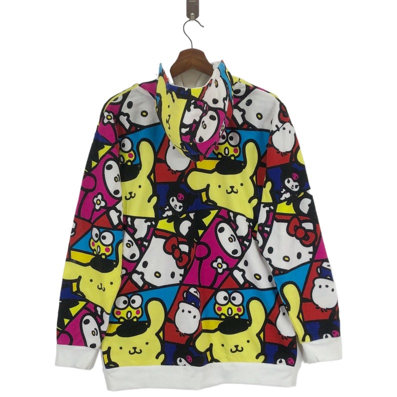 Op de afbeelding: Een wit hoodie met een kleurrijke all-over print met cartoonfiguren. De print bevat de kleuren geel, roze, blauw, rood, zwart en groen. De personages zijn een konijn, een kat en een kikker.