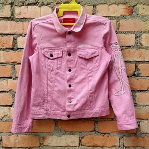 May include: A pink denim jacket with a white graphic on the right sleeve. The jacket has a button closure and two pockets on the front.