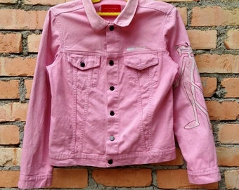 May include: A pink denim jacket with a white graphic on the right sleeve. The jacket has a button closure and two pockets on the front.