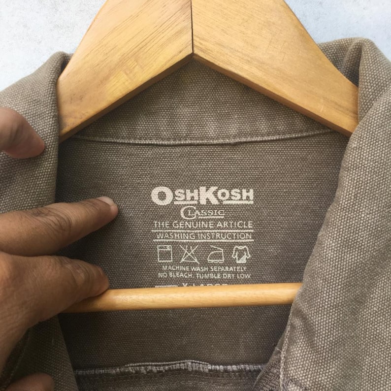 K&ouml;nnte beinhalten: Ein braunes OshKosh Classic-Etikett mit wei&szlig;em Text, der "The Genuine Article Washing Instruction" lautet und ein Waschanleitungssymbol enth&auml;lt. Das Etikett ist an einem braunen Kleidungsst&uuml;ck befestigt.