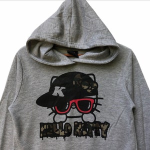 May include: Gray hooded sweatshirt with a black and white Hello Kitty graphic wearing a black and camo baseball cap and red sunglasses. The text "Hello Kitty" is written in black with a camo pattern.