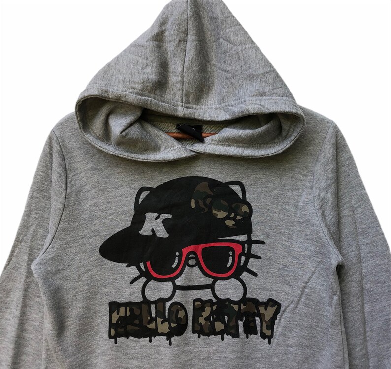 May include: Gray hooded sweatshirt with a black and white Hello Kitty graphic wearing a black and camo baseball cap and red sunglasses. The text "Hello Kitty" is written in black with a camo pattern.