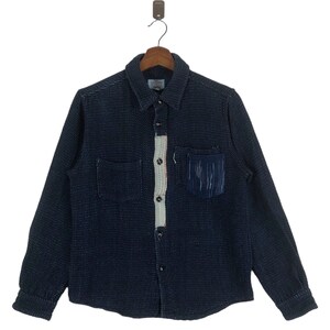May include: A dark blue long-sleeved shirt with a white and cream patterned panel down the front. The shirt has a button-up closure and two pockets on the front. The pocket on the left side has a blue and white fringe detail.