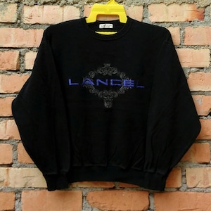 Könnte beinhalten: Schwarzer Pullover mit einem blauen gestickten Logo, auf dem in einer dekorativen Schrift "LANCEL" steht.