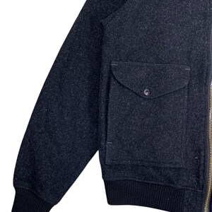 Puede incluir: Chaqueta bomber de lana gris oscuro con cuello y pu&ntilde;os de canal&eacute; negros. La chaqueta presenta un &uacute;nico bolsillo con solapa y cierre de bot&oacute;n y una cremallera dorada.