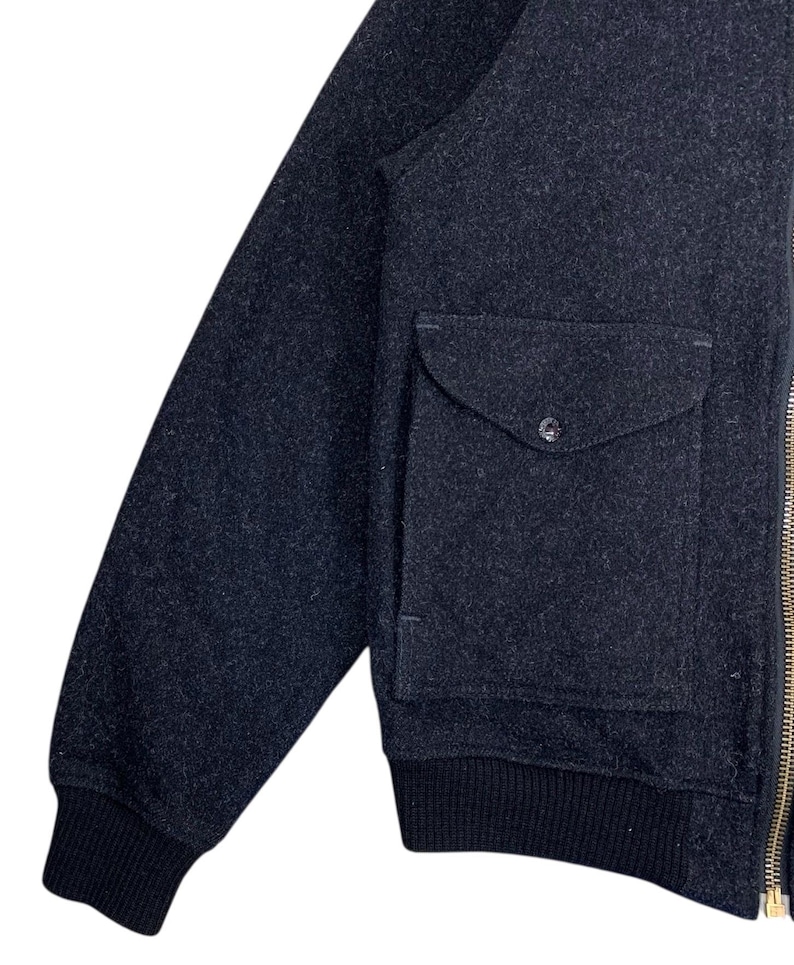 Puede incluir: Chaqueta bomber de lana gris oscuro con cuello y pu&ntilde;os de canal&eacute; negros. La chaqueta presenta un &uacute;nico bolsillo con solapa y cierre de bot&oacute;n y una cremallera dorada.