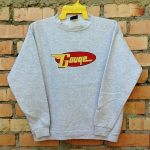 Könnte beinhalten: Ein grauer Pullover mit einem roten und gelben Logo, auf dem "Gouge" mit einem Flügel auf jeder Seite des Wortes steht.