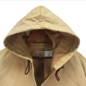 May include: A tan Carhartt hooded jacket with a zipper closure and a brown drawstring. The jacket has a brown label with the Carhartt logo on the front.