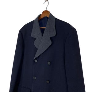 Puede incluir: Un abrigo azul marino de doble botonadura con un cuello de dise&ntilde;o gris y negro en contraste. El abrigo tiene cuatro botones negros y dos bolsillos delanteros. El abrigo est&aacute; colgado en una percha de madera.