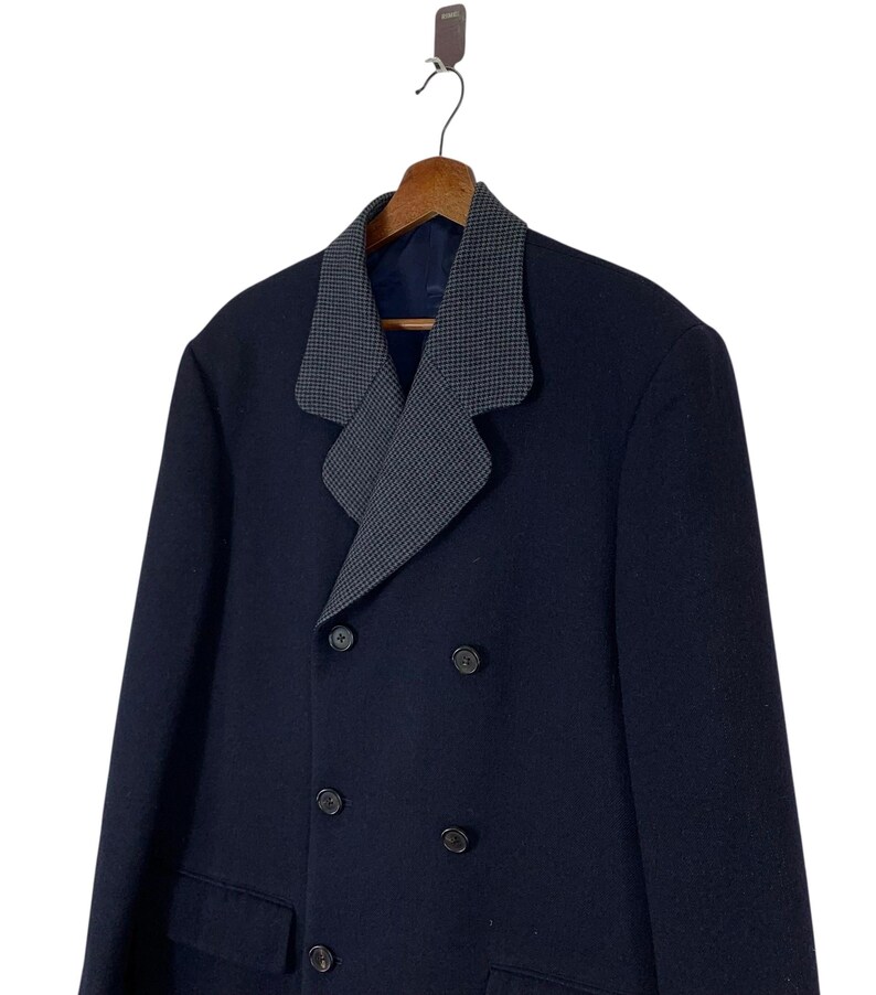 Puede incluir: Un abrigo azul marino de doble botonadura con un cuello de dise&ntilde;o gris y negro en contraste. El abrigo tiene cuatro botones negros y dos bolsillos delanteros. El abrigo est&aacute; colgado en una percha de madera.
