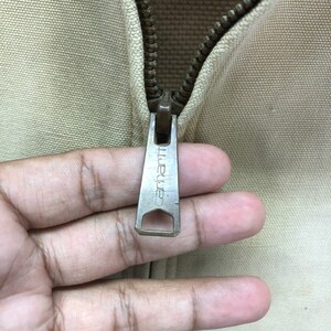 May include: Close-up of a zipper pull on a beige jacket. The zipper pull is metal and has the word "Carhartt" engraved on it.