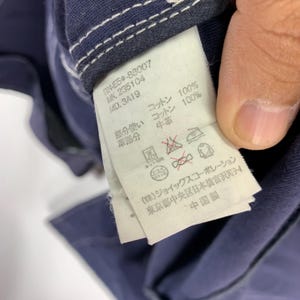 May include: A dark blue garment featuring a white interior care label with black printed text. The label displays product codes, material composition including 100% cotton, and various laundry instruction symbols. The fabric has white stitching.