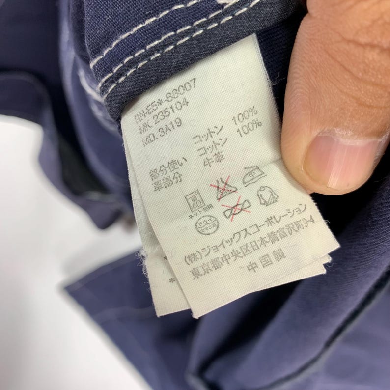 May include: A dark blue garment featuring a white interior care label with black printed text. The label displays product codes, material composition including 100% cotton, and various laundry instruction symbols. The fabric has white stitching.
