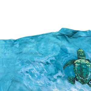 May include: A turquoise tie-dye t-shirt featuring a detailed illustration of a green sea turtle swimming in the ocean. The design includes white wave patterns, creating a dynamic and aquatic theme. The shirt's fabric has a soft, textured appearance.