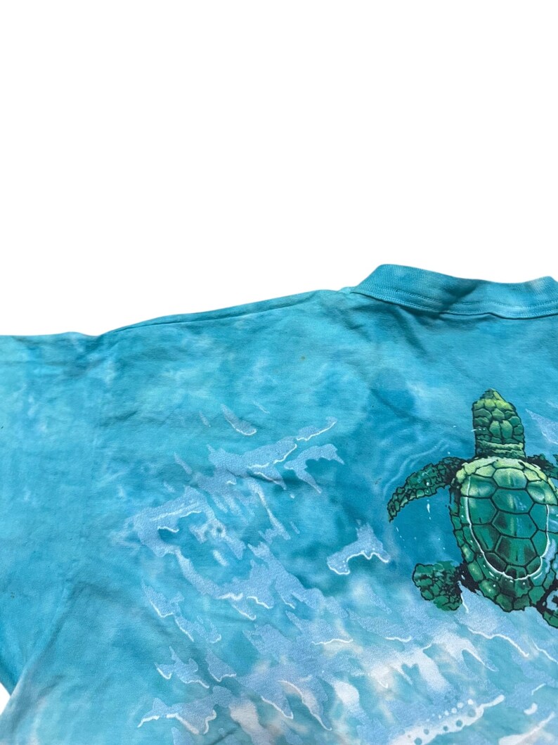 May include: A turquoise tie-dye t-shirt featuring a detailed illustration of a green sea turtle swimming in the ocean. The design includes white wave patterns, creating a dynamic and aquatic theme. The shirt's fabric has a soft, textured appearance.