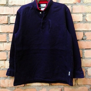 Puede incluir: Una camisa de manga larga azul oscuro con cuello abotonado y un pequeño bolsillo en el lado izquierdo. La camisa tiene un ribete rojo y blanco alrededor del cuello.