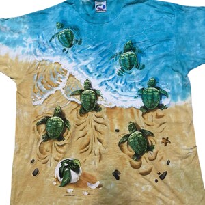 May include: A blue and tan t-shirt featuring a beach scene with sea turtles. The shirt has a tie-dye effect with a blue ocean and tan sand. Several green sea turtles are depicted on the shirt, some swimming in the water and others on the sand.