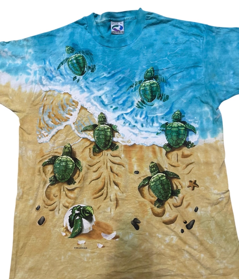 May include: A blue and tan t-shirt featuring a beach scene with sea turtles. The shirt has a tie-dye effect with a blue ocean and tan sand. Several green sea turtles are depicted on the shirt, some swimming in the water and others on the sand.
