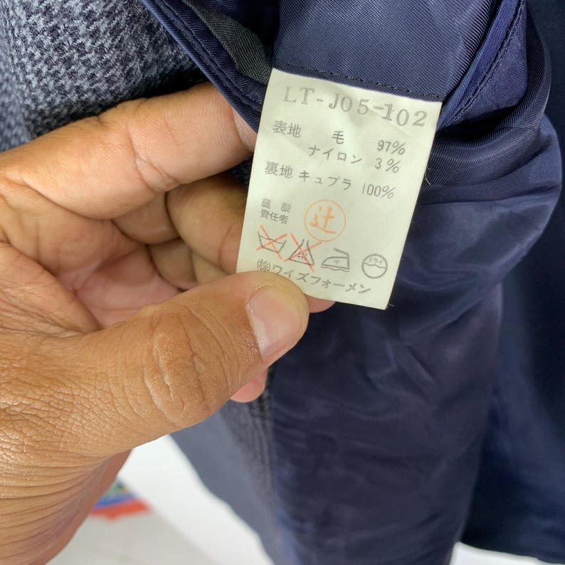 Puede incluir: Primer plano de una etiqueta de prenda con el texto "LT-J05-102". La etiqueta enumera la composici&oacute;n de la tela: 97% lana, 3% nailon y 100% forro de cupro. Tambi&eacute;n se incluyen s&iacute;mbolos de cuidado y texto japon&eacute;s.