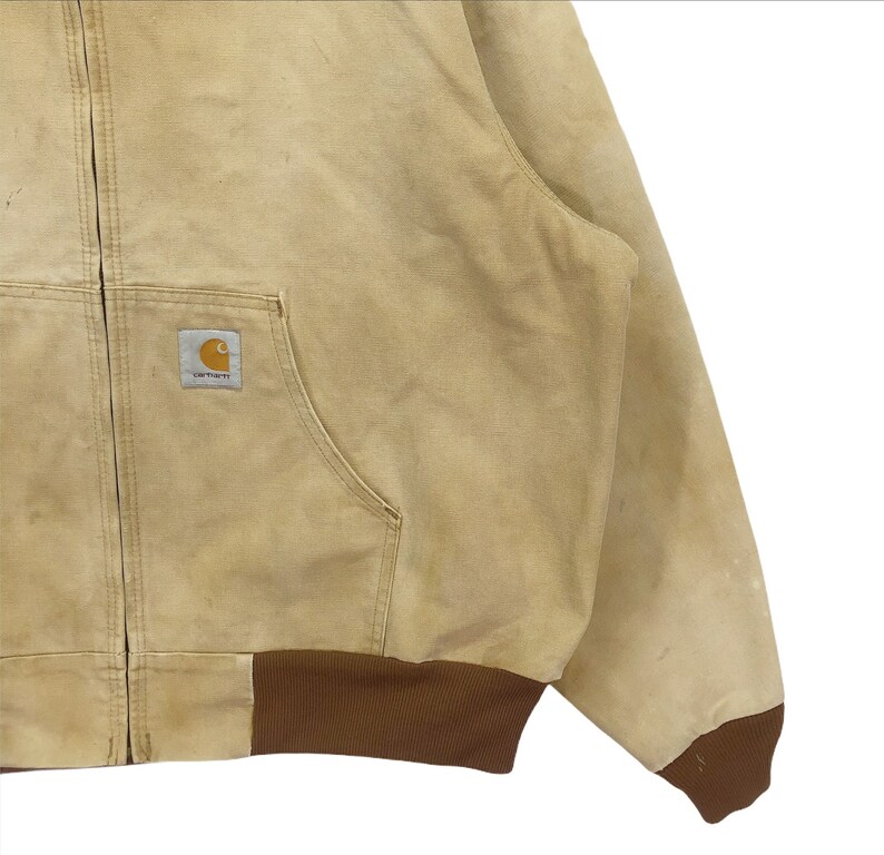 May include: A tan Carhartt work jacket with a brown ribbed waistband and cuffs. The jacket has a zipper closure and a patch pocket on the left chest with the Carhartt logo.