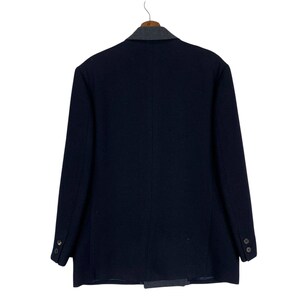 Puede incluir: Un blazer azul marino con un cuello y solapa estampados en gris y blanco contrastantes. La parte trasera de la chaqueta es visible, colgada de una percha de madera. El blazer tiene dos botones en cada pu&ntilde;o y una sola abertura.