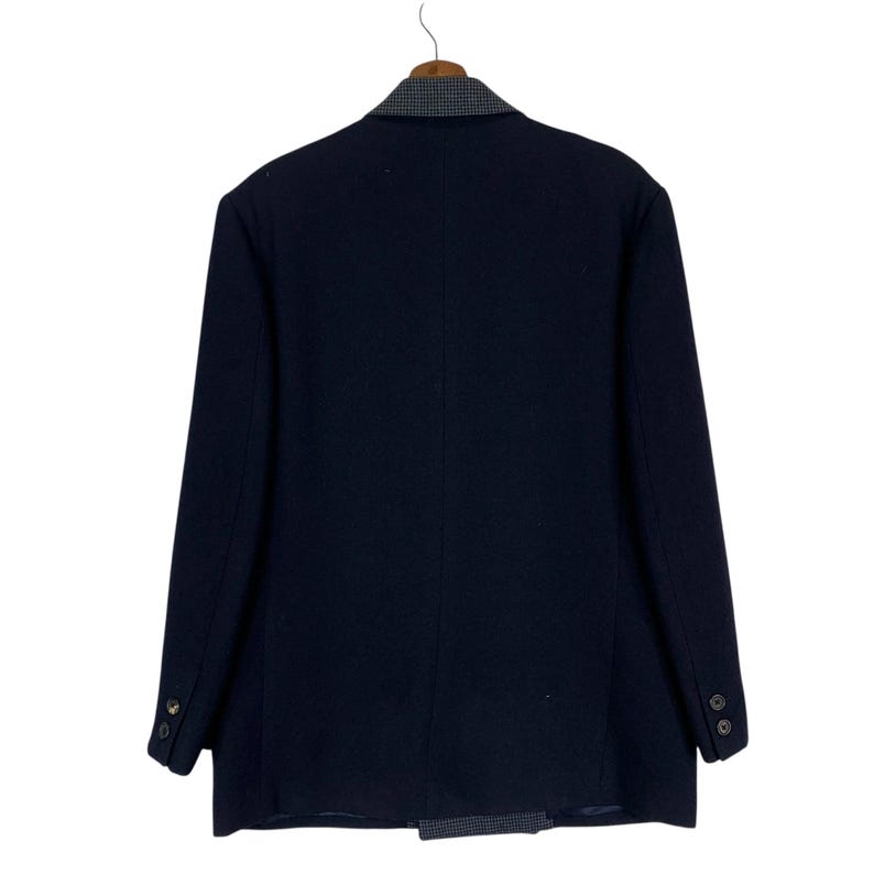 Puede incluir: Un blazer azul marino con un cuello y solapa estampados en gris y blanco contrastantes. La parte trasera de la chaqueta es visible, colgada de una percha de madera. El blazer tiene dos botones en cada pu&ntilde;o y una sola abertura.