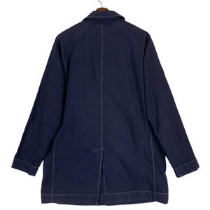 May include: A dark navy blue jacket featuring long sleeves, a standard collar, and prominent white contrast stitching along the seams. The back of the jacket displays a central vertical seam and a single vent at the bottom hem, hanging on a hanger.