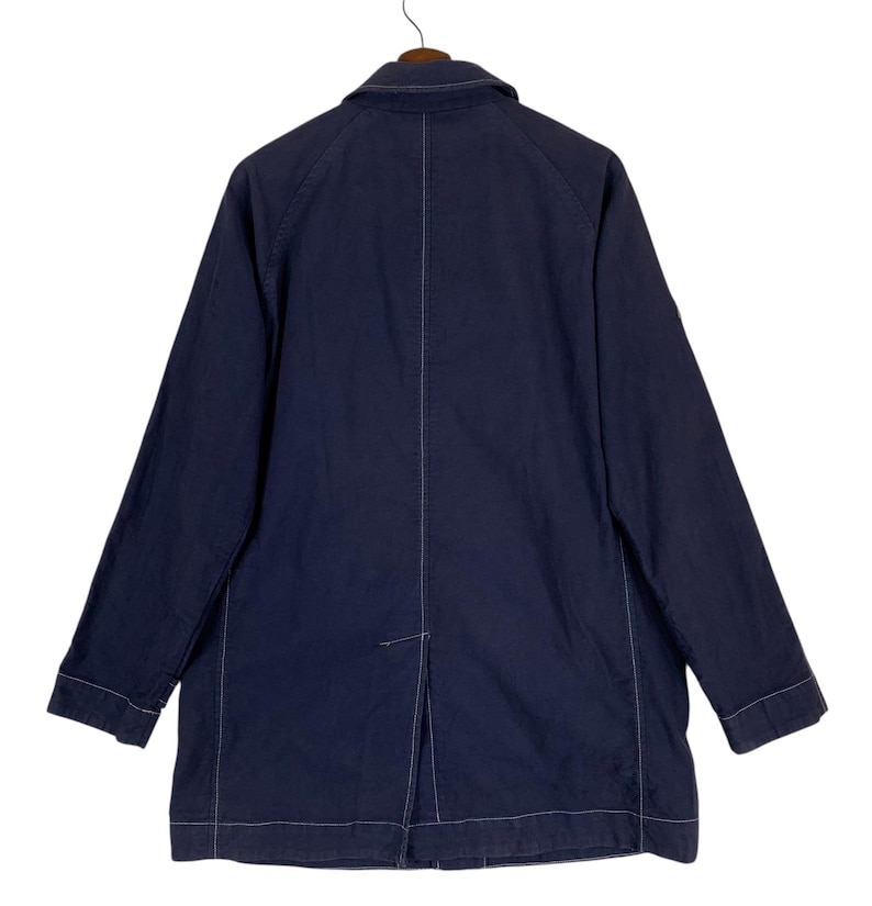 May include: A dark navy blue jacket featuring long sleeves, a standard collar, and prominent white contrast stitching along the seams. The back of the jacket displays a central vertical seam and a single vent at the bottom hem, hanging on a hanger.