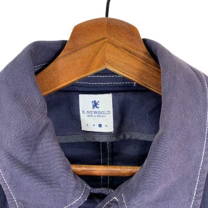 May include: A dark navy blue jacket with a wide collar and contrasting white stitching. The interior neck label features a blue lion logo, the text R.NEWBOLD BORN IN BRITAIN, and size indicators with the letter L highlighted in a blue circle.