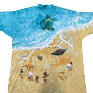 May include: A tie-dye t-shirt featuring a beach scene with a sea turtle, seashells, and a bird. The top of the shirt has a blue ocean design, transitioning to a sandy beach at the bottom. The shirt is a unique, artistic design.