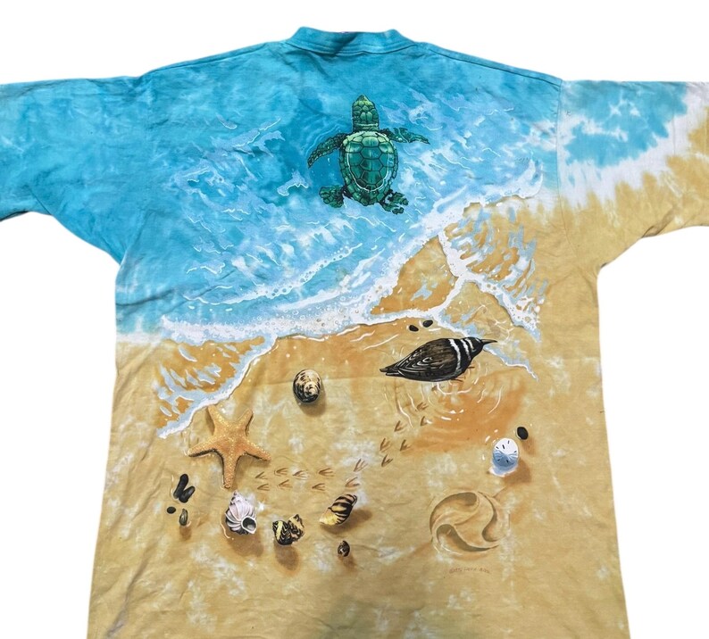 May include: A tie-dye t-shirt featuring a beach scene with a sea turtle, seashells, and a bird. The top of the shirt has a blue ocean design, transitioning to a sandy beach at the bottom. The shirt is a unique, artistic design.