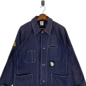 May include: A dark blue long-sleeve jacket featuring white contrast stitching, a classic collar, and a button-front closure. The garment includes multiple patch pockets on the front, a small yellow triangular patch on the sleeve, and a dark green star-shaped patch on the chest.