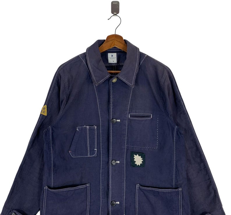 May include: A dark blue long-sleeve jacket featuring white contrast stitching, a classic collar, and a button-front closure. The garment includes multiple patch pockets on the front, a small yellow triangular patch on the sleeve, and a dark green star-shaped patch on the chest.