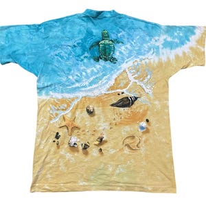 May include: A tie-dye t-shirt featuring a beach scene with a sea turtle, bird, starfish, and seashells. The top of the shirt has a blue and white ocean design, transitioning to a sandy beach with detailed graphics.