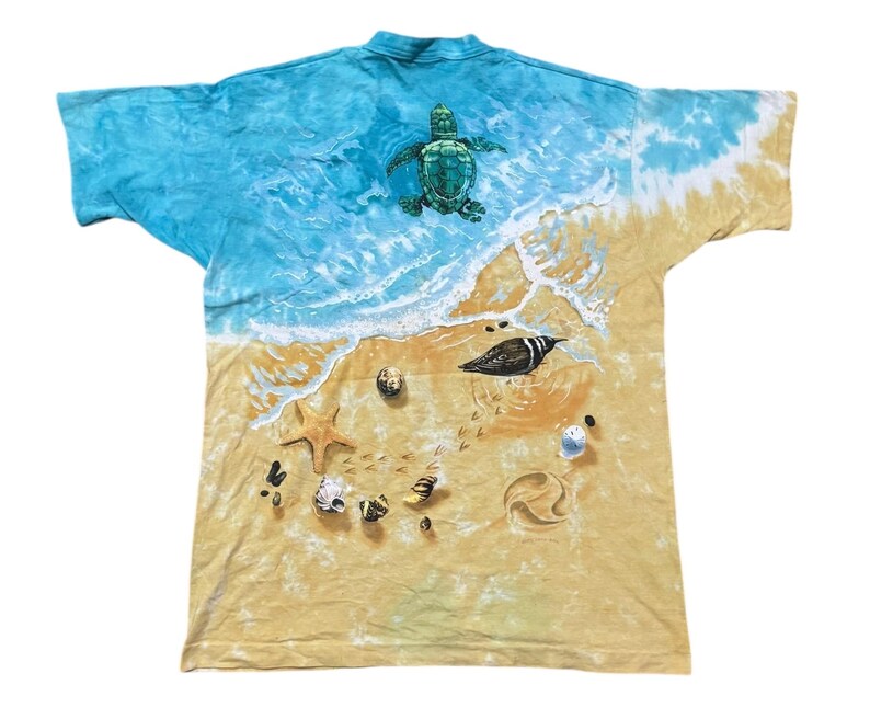 May include: A tie-dye t-shirt featuring a beach scene with a sea turtle, bird, starfish, and seashells. The top of the shirt has a blue and white ocean design, transitioning to a sandy beach with detailed graphics.