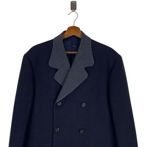 Puede incluir: Un abrigo azul marino de doble botonadura con un cuello de dise&ntilde;o gris y negro en contraste. El abrigo tiene dos filas de botones y est&aacute; colgado de una percha de madera.