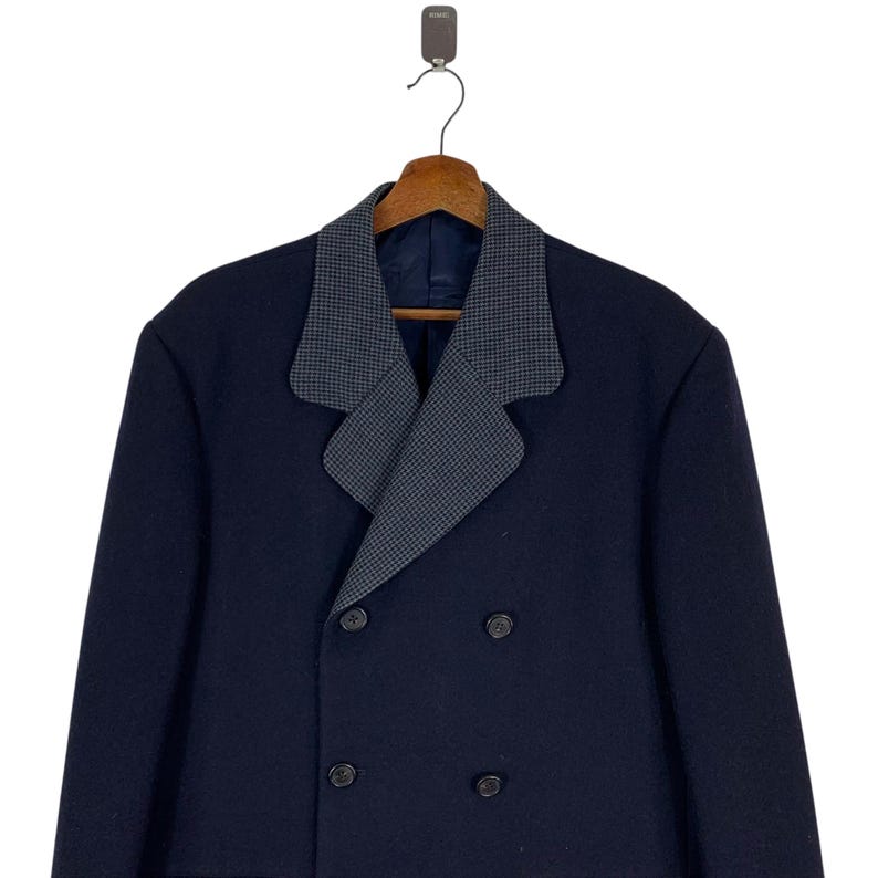 Puede incluir: Un abrigo azul marino de doble botonadura con un cuello de dise&ntilde;o gris y negro en contraste. El abrigo tiene dos filas de botones y est&aacute; colgado de una percha de madera.