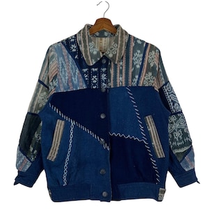 May include: A long-sleeve jacket featuring a patchwork design of solid navy blue panels and patterned sections in shades of blue, grey, and white. The garment includes a collared neckline, a front button closure, and white decorative stitching.