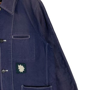 May include: A dark blue long-sleeve jacket featuring a pointed collar, white contrast stitching, and dark buttons. The front includes a chest pocket and a lower pocket, with a small square dark green patch displaying a white star-shaped graphic.
